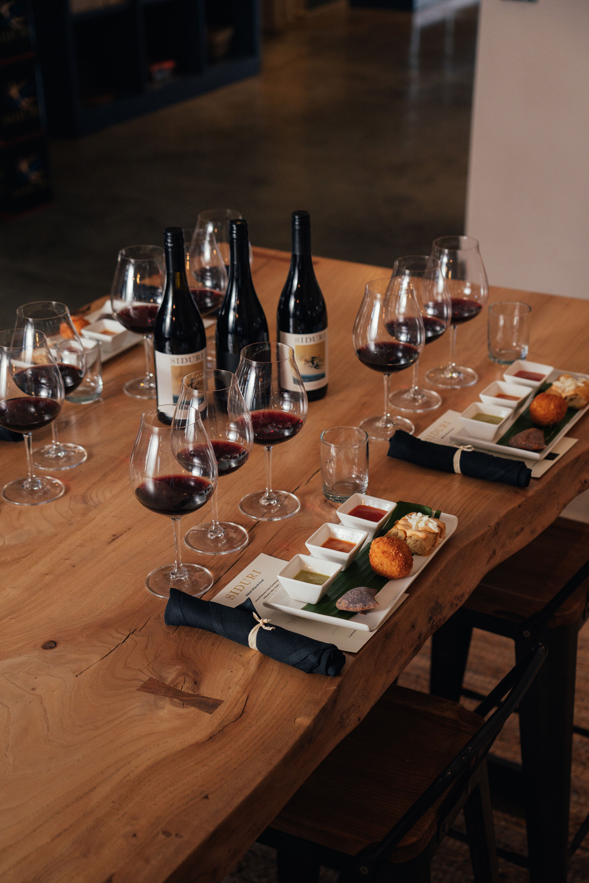 Four plates of food on a wood table with three bottles of wine and wine glasses