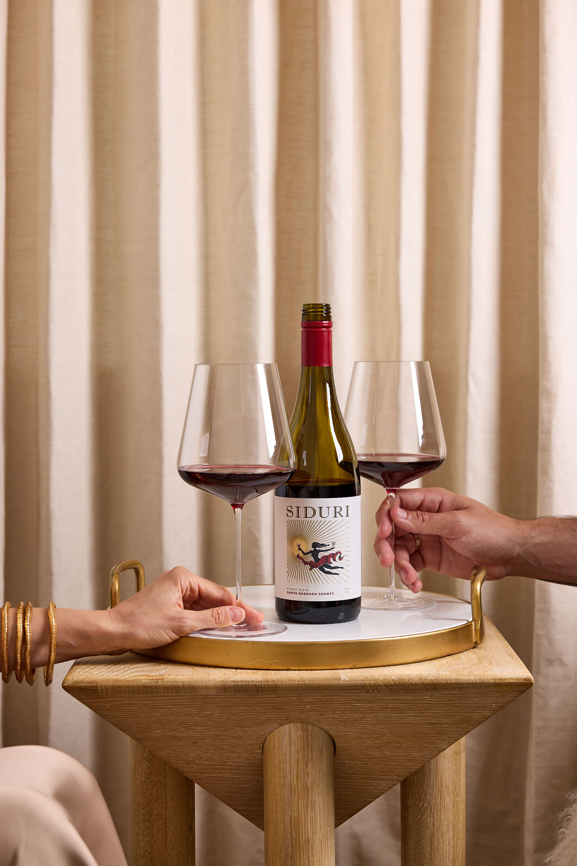 A bottle of red wine on a wood table with beige curtains in the background, and two people's hands holding wine glasses A bottle of red wine on a wood table with beige curtains in the background, and two people's hands holding wine glasses